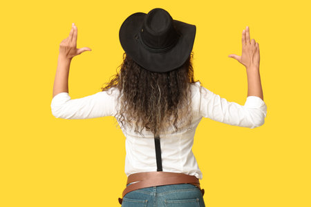 Beautiful African-American cowgirl making guns with her hands on yellow background, back viewの写真素材