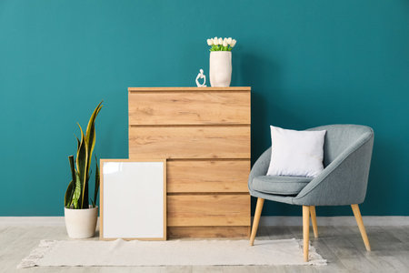 Armchair, chest of drawers, houseplant and frame near blue wall in room