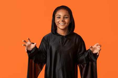Cute happy African-American girl dressed for Halloween party on orange background