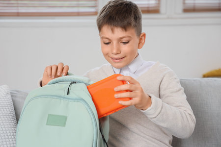 Little boy putting school lunchbox into backpack on sofa at home