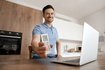 Handsome man with air conditioner remote control and laptop in kitchen, closeupの写真素材