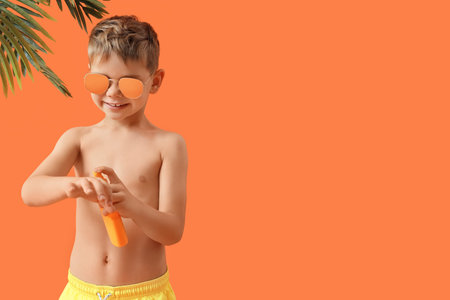 Cute little boy in sunglasses applying sunscreen cream and palm leaves on orange background