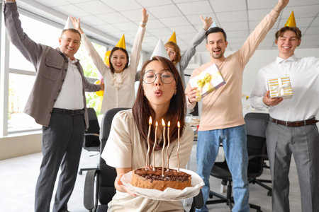 Young Asian businesswoman blowing out candles on cake at birthday party in office