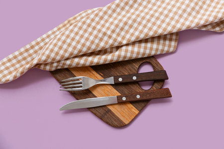 Table setting with checkered napkin, cutlery and wooden board on purple background