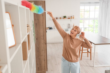Mature woman in headphones with pp-duster cleaning shelf at homeの写真素材
