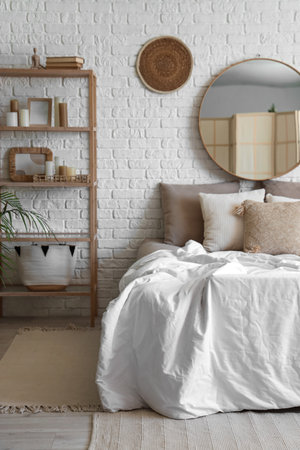 Comfortable bed with pillows and mirror on white brick wall in stylish bedroomの写真素材