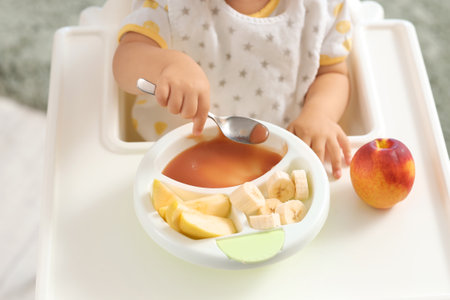 Cute little baby eating tasty food and fruits on chair in kitchen at home, closeupの写真素材