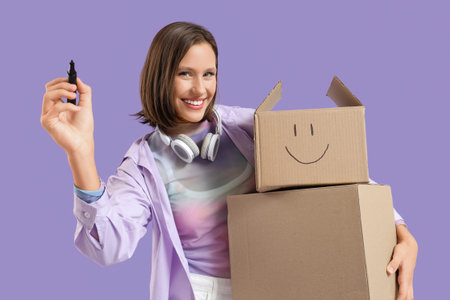 Beautiful young happy woman with headphones drawing smile face on parcel box against purple backgroundの写真素材