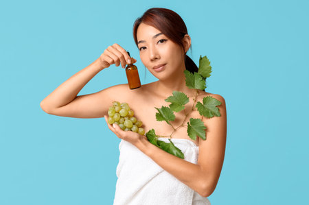 Beautiful young Asian woman with bottle of cosmetic grape seed oil and leaves on blue backgroundの写真素材