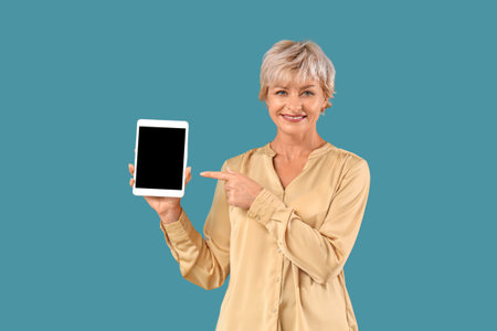 Beautiful mature woman pointing at blank tablet computer on blue background
