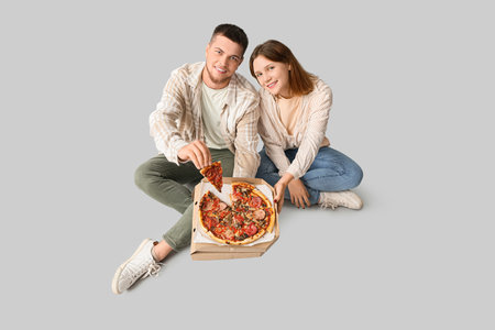 Beautiful young happy couple with cardboard box of tasty pizza sitting on gray backgroundの写真素材
