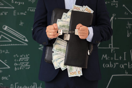Handcuffed male teacher with briefcase and bribe near blackboard, closeup. Corruption conceptの写真素材