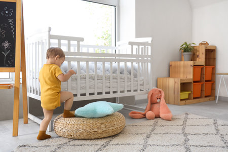 Cute baby girl climbing onto pouf near crib at homeの写真素材