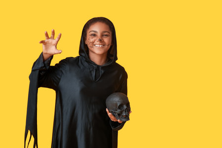 Cute happy African-American girl with human skull dressed for Halloween party on yellow backgroundの写真素材