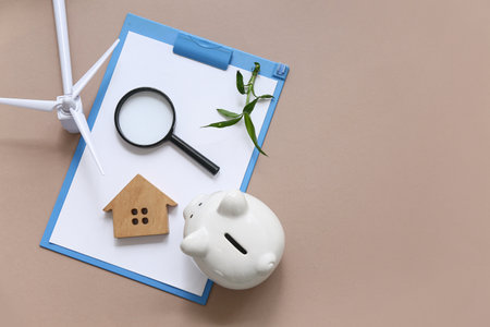 Clipboard with plant, magnifier, house figure, piggy bank and wind turbine on brown background. Green Energy conceptの写真素材