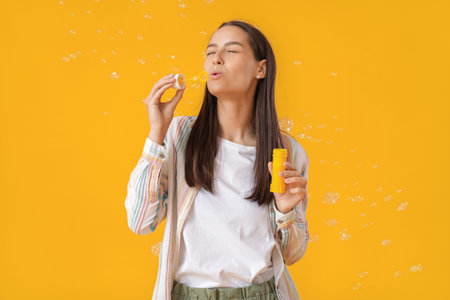 Young woman blowing soap bubbles on yellow background