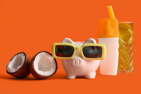 Piggy bank, sunglasses, cocktail, sunscreen and coconut on orange backgroundの写真素材