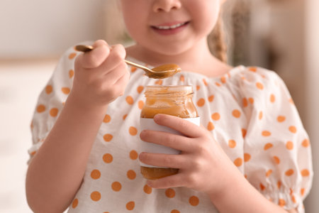 Cute little girl with spoon and jar of jam at homeの写真素材
