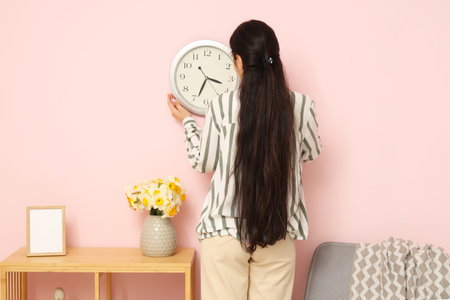 Young woman hanging clock on pink wall near shelving unit in beautiful room