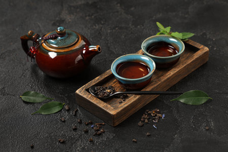 Teapot with cups, spoon of dry tea and leaves on black grunge backgroundの写真素材