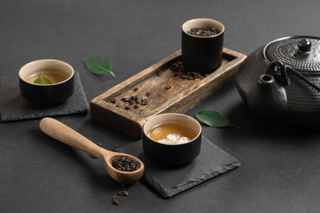 Teapot with cups, wooden spoon of dry tea and leaves on black background