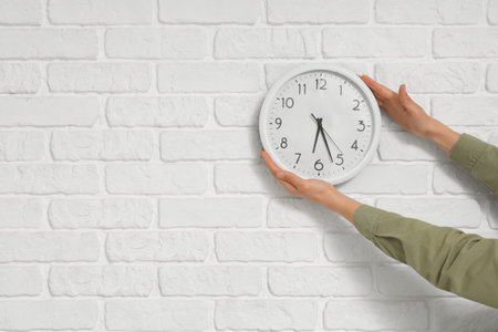 Young woman hanging clock on white brick wall in room