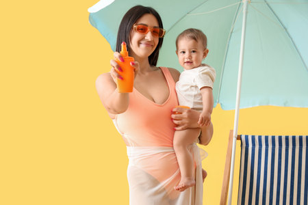 Young mother with her little baby applying sunscreen cream and beach umbrella on yellow backgroundの写真素材
