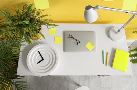 Stylish workplace with laptop, clock and lamp near yellow wall