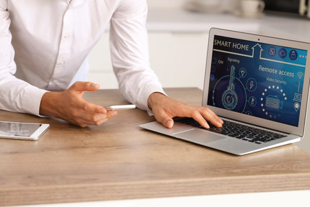Young man using mobile phone and laptop with smart home application at table in kitchen, closeup