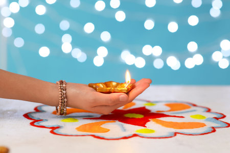 Female hand with diya lamp for Diwali and colorful powder on white table against blue blurred...