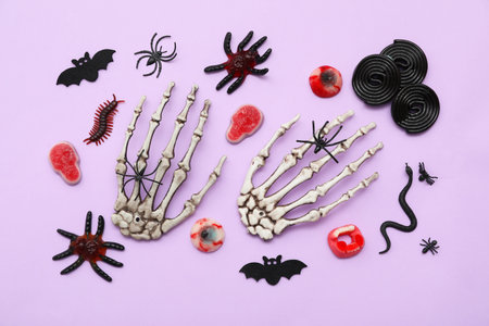 Composition with skeleton hands and different candies for Halloween on lilac background