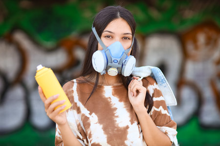 Young woman in respirator with spray paint can against graffiti wall outdoorsの写真素材
