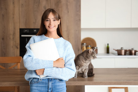 Beautiful young happy woman with laptop and cute cat on table in kitchenの写真素材