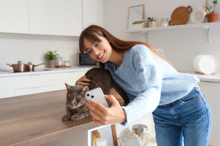 Beautiful young happy woman with cute cat and mobile phone taking selfie in kitchen at homeの写真素材