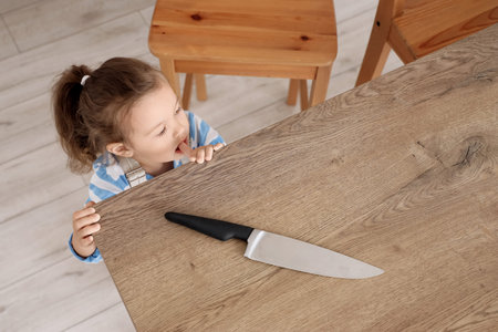 Cute little girl taking knife from table in kitchen, top view. Child at risk