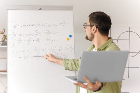 Young male math teacher with laptop and formulas on whiteboard giving online lesson at home
