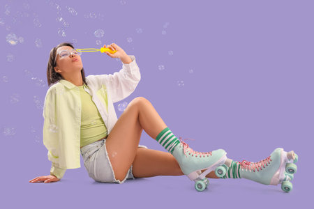 Young woman in roller skates sitting and blowing soap bubbles on lilac background