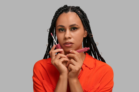 Young African-American woman with braids and beautiful makeup holding lip gloss on gray background