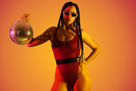 Beautiful young African-American woman with braids holding disco ball on color background