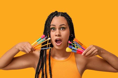 Shocked young African-American woman with braids and beautiful makeup holding brushes on yellow...