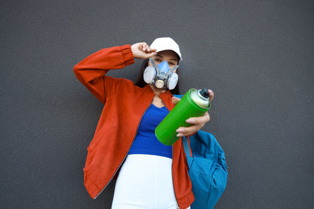 Young woman in respirator with spray paint can and backpack near building wall on streetの写真素材