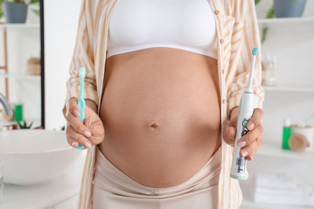 Young pregnant woman holding plastic and electric tooth brushes in white bathroomの写真素材