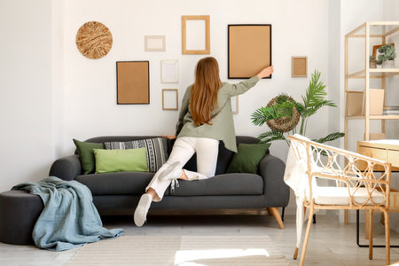 Young woman hanging picture on white wall near sofa in living roomの写真素材