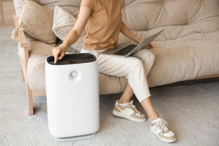 Young woman sitting on sofa and turning on modern humidifier in beautiful living room, closeupの写真素材