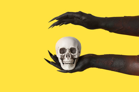Black hands of witch with human skull on yellow background. Halloween celebration