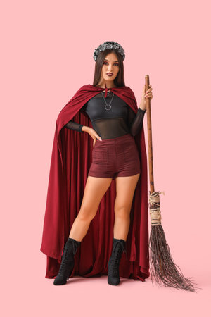 Beautiful young Asian woman dressed as witch for Halloween with magic broom on pink background