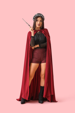 Beautiful young thoughtful Asian woman dressed as witch for Halloween with magic wand on pink backgroundの写真素材