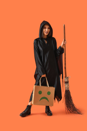 Beautiful young happy Asian woman dressed as witch for Halloween with shopping bag and magic broom on orange backgroundの写真素材