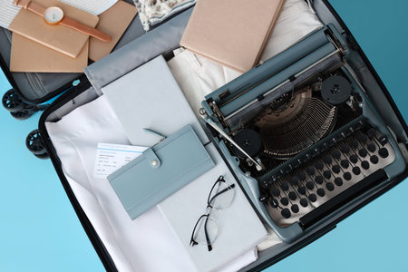 Open suitcase with vintage typewriter, books and clothes on blue background, top view