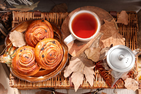 Teapot with cup of hot beverage, tasty buns and autumn leaves on wicker basketの写真素材
